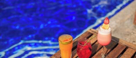 Deux boissons sur une table en bois, près d’une piscine à l’eau bleue.