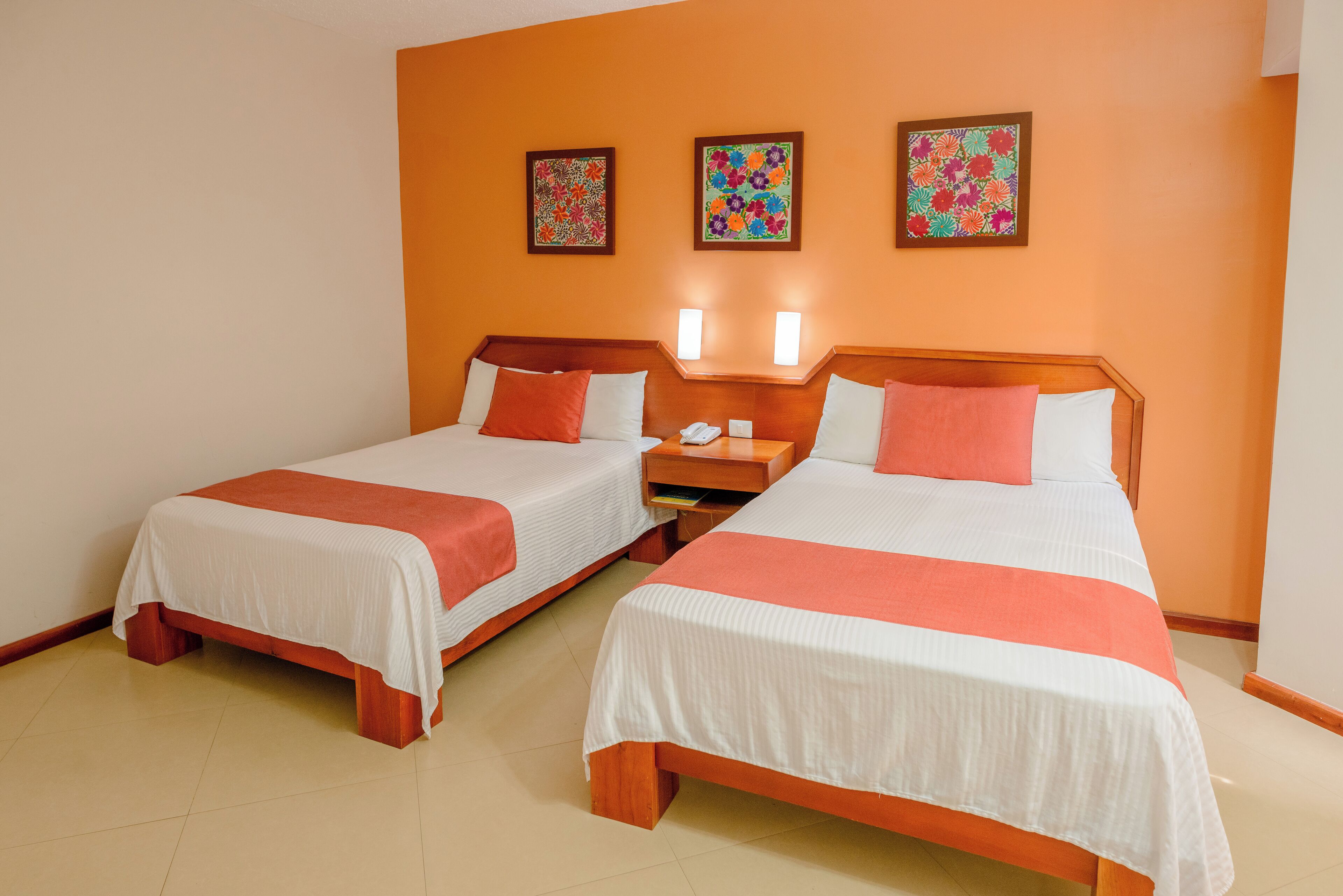 Une chambre d’hôtel avec deux lits, chacun agrémenté de linge de lit blanc et de touches de couleur orange, ainsi que trois œuvres d’art murales aux motifs floraux.