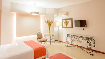 A hotel room with two beds, a chair, a table with a vase of flowers, a lamp, a TV, and a decorative metal bench.