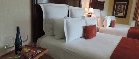 A hotel room with two beds, a wooden headboard, white bedding, a tray with fruit and a bottle, and a lamp.