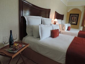 Habitación de hotel con dos camas, cabecero de madera, ropa de cama blanca, una bandeja con fruta y una botella, y una lámpara.
