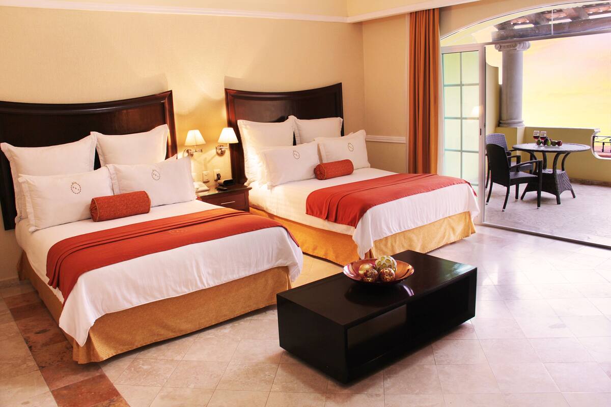 Une chambre d’hôtel avec deux lits, une table de chevet avec des lampes, une table basse ornée d’un vase décoratif et un balcon avec une table et des chaises.