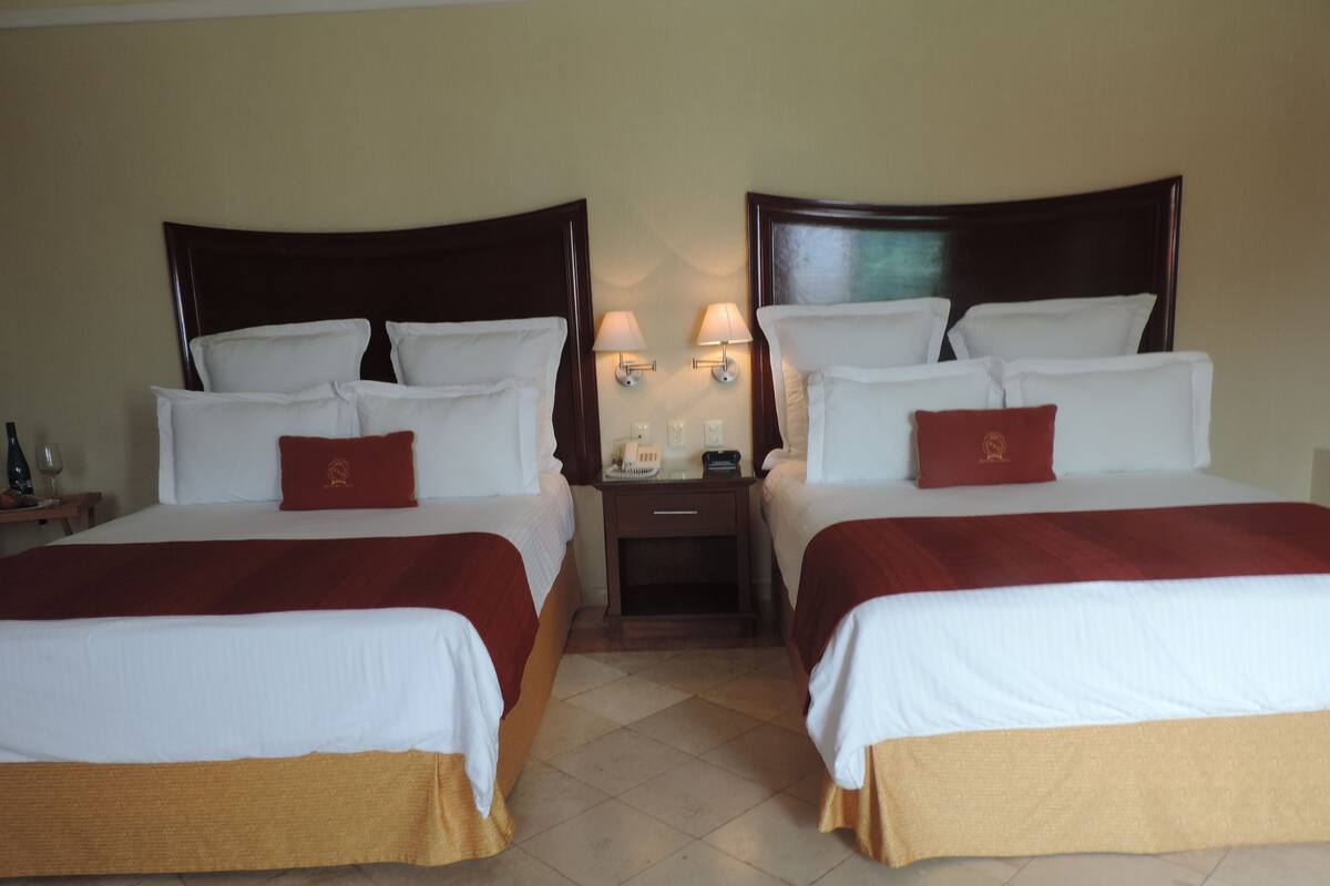 Une chambre d’hôtel avec deux lits, chacun agrémenté de draps blancs et d’oreillers rouges, une table de chevet avec une lampe et une petite table sur laquelle se trouvent une bouteille et un verre.