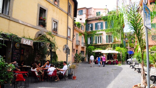 An outdoor café with tables and chairs, surrounded by buildings with balconies and potted plants. - Little Rhome Suites (Rome)