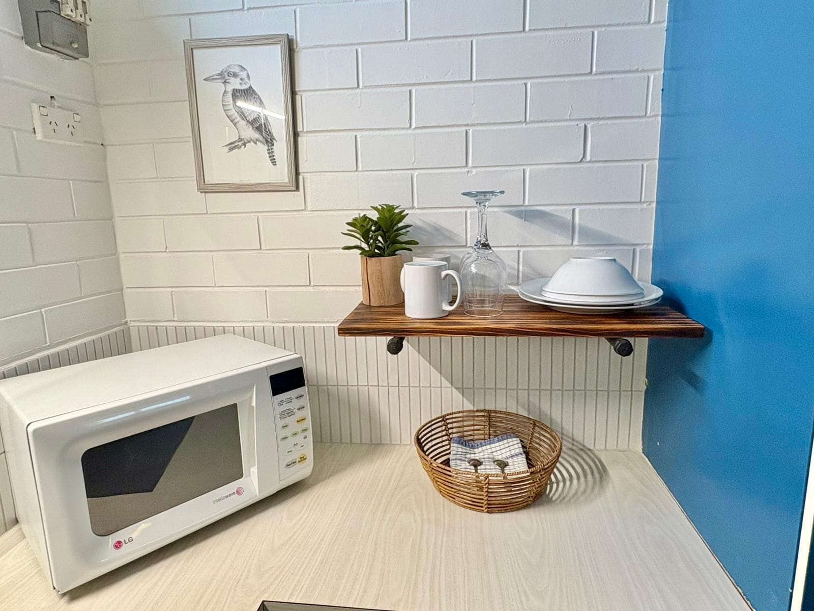 A kitchen with a white microwave, a wooden shelf with a plant, a glass pitcher, and white dishes.