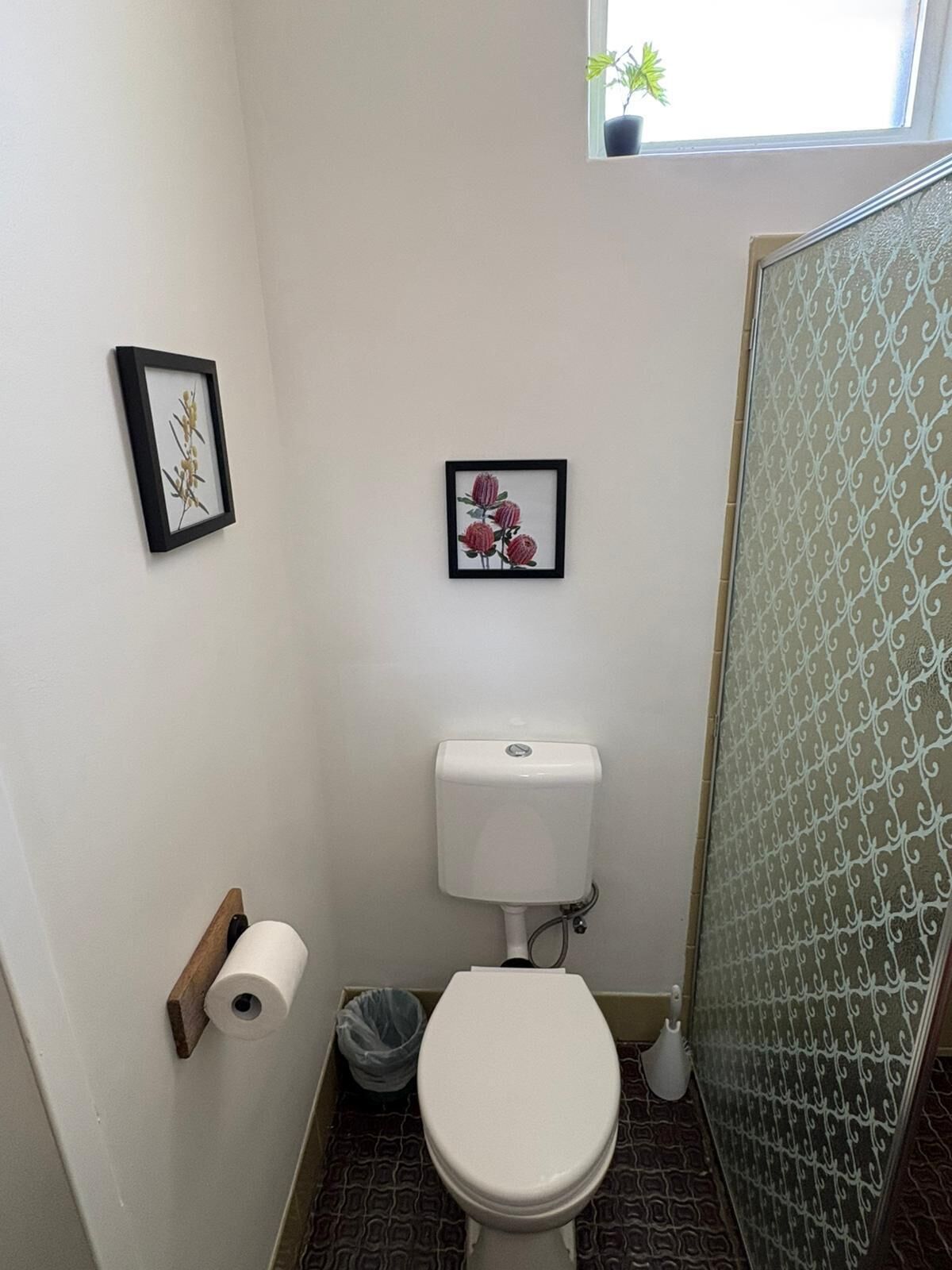 A small bathroom with a toilet, a framed picture on the wall, and a window with a plant.
