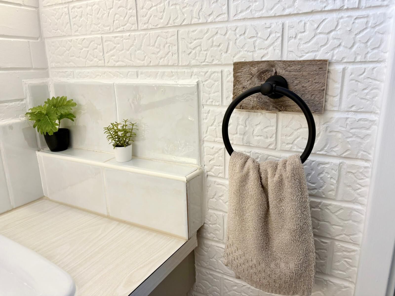 Un baño con azulejos blancos con textura, un perchero de madera y dos plantas en macetas sobre una repisa.