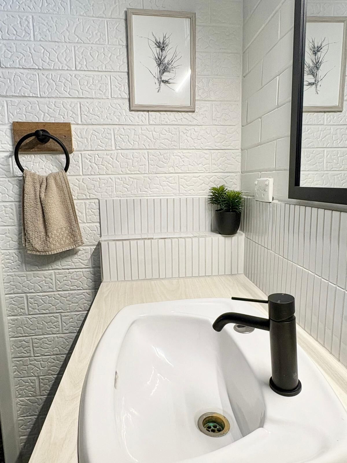 Ein Badezimmer mit einem weißen Waschbecken, einem schwarzen Wasserhahn und einem Handtuch, das an einem Metallring hängt. An der Wand befindet sich eine gerahmte Pflanze und auf der Ablage ein kleiner Blumentopf.