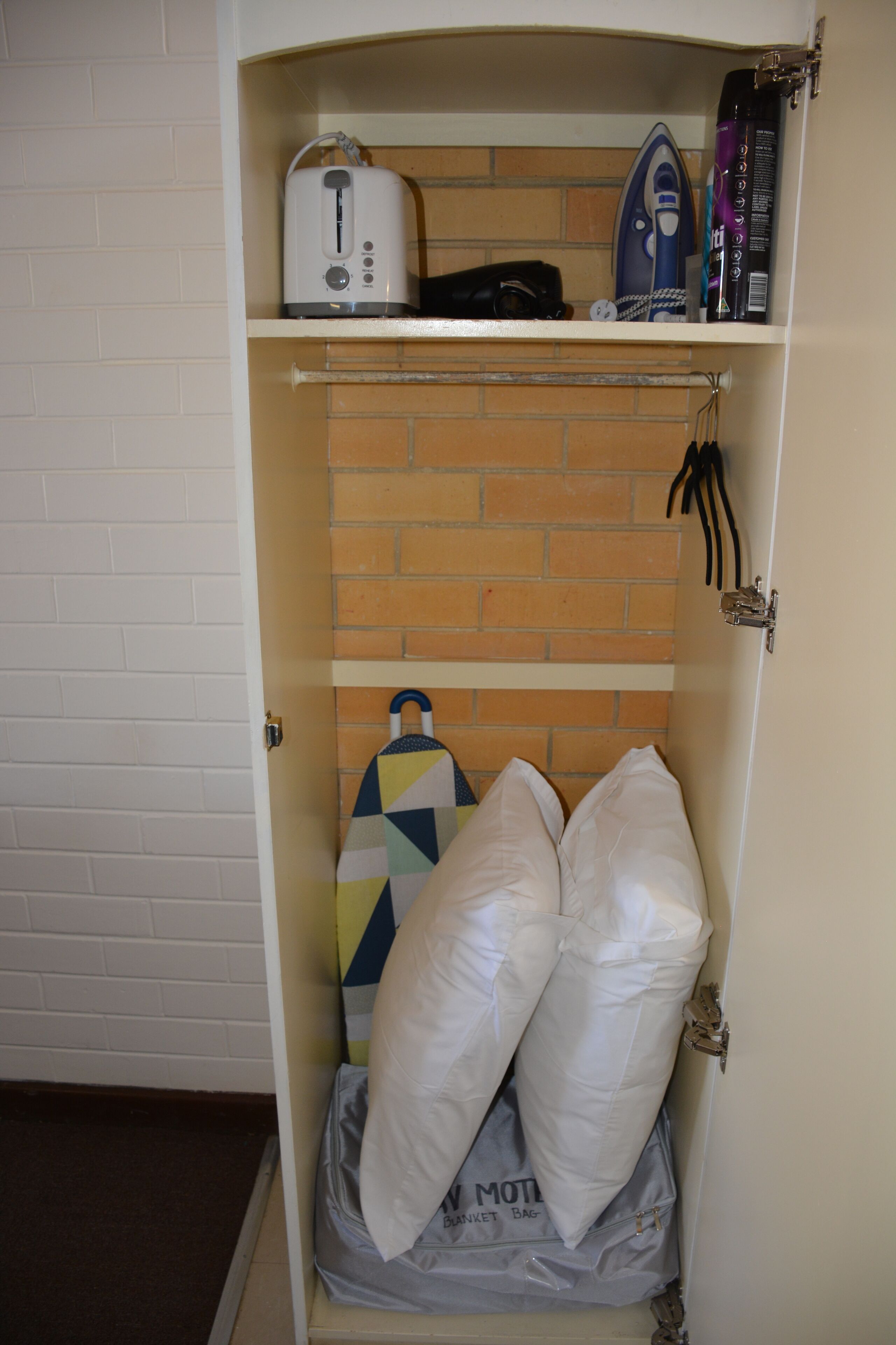 A small storage space with a brick-patterned wall, containing a toaster, an iron, and a bag labeled "MOTEL".