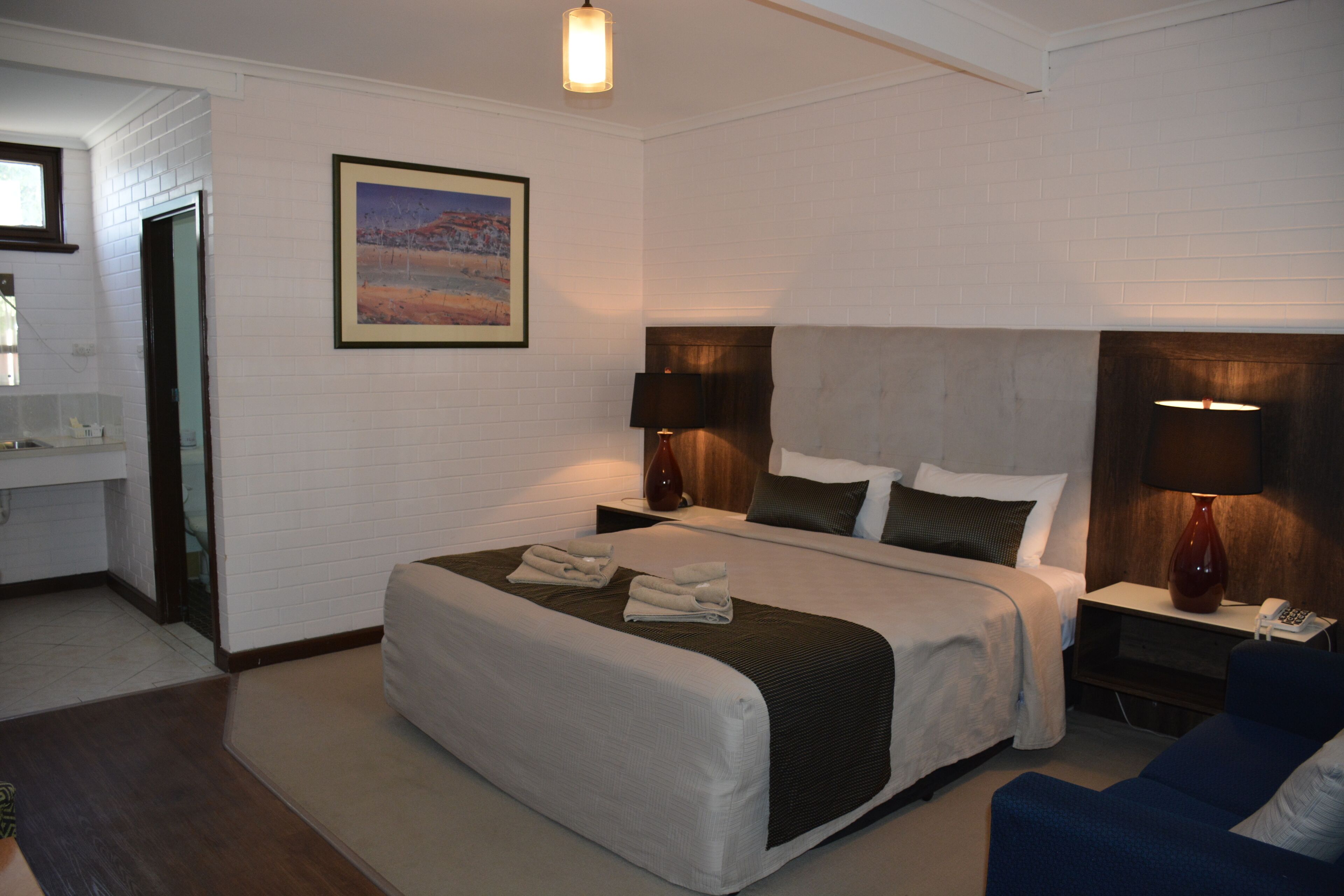 A hotel room with a large bed, a bedside table with a lamp, a small desk with a telephone, and a painting on the wall.