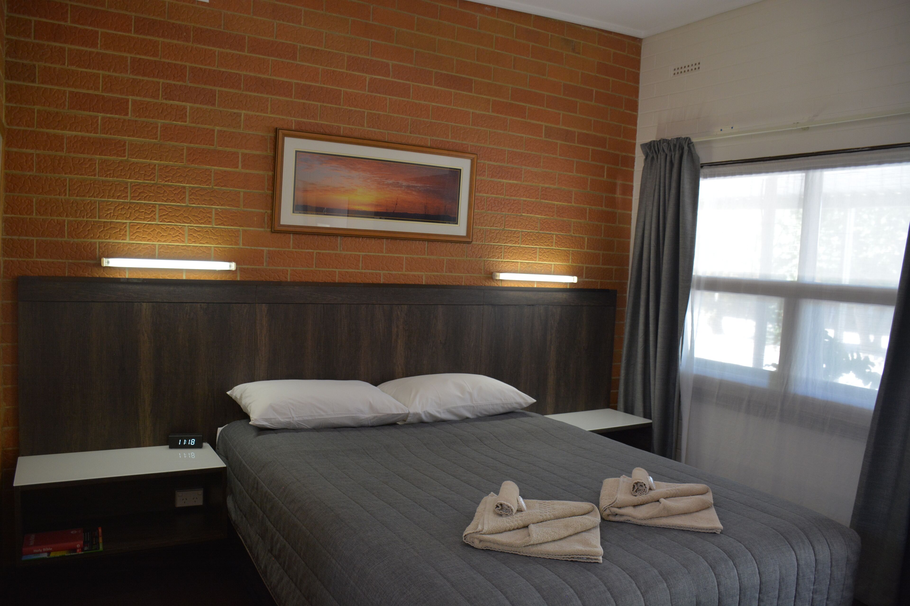 A hotel room with a brick wall, a painting, a bed with pillows, a bedside table, and a window with curtains.