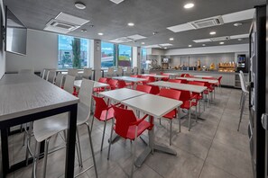 Une cafétéria moderne avec des tables et des chaises blanches et rouges, de grandes fenêtres et un espace cuisine.