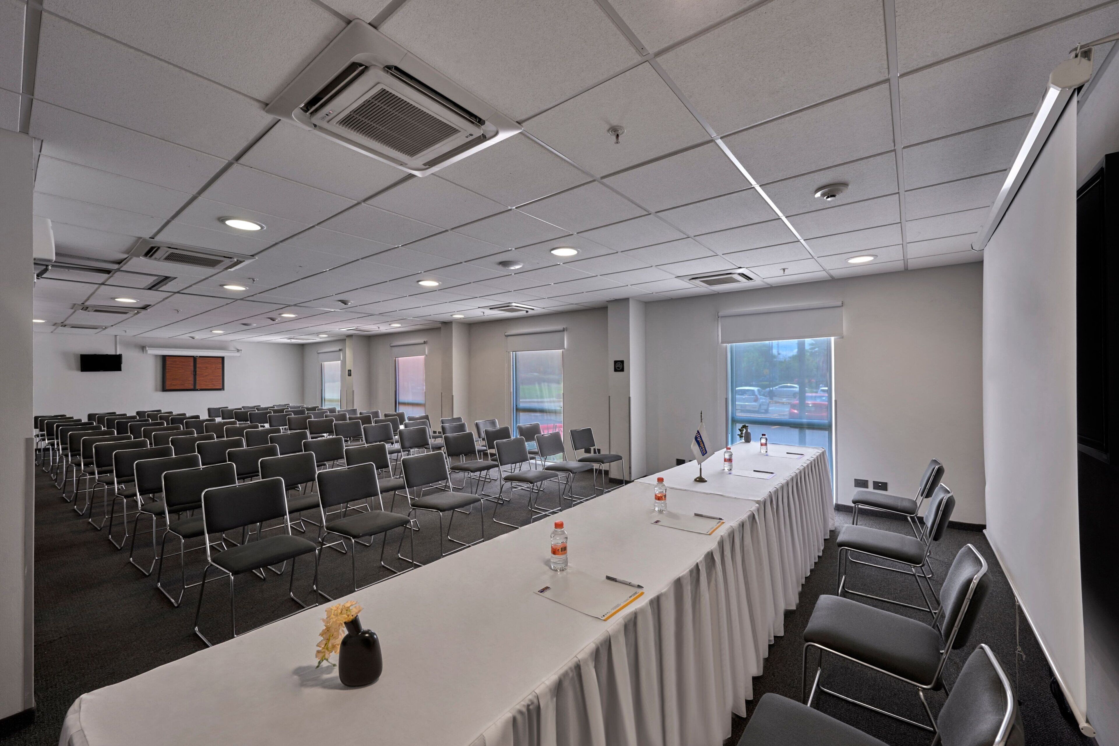 Un salón de conferencias con filas de sillas, una larga mesa con botellas de agua y un amplio ventanal con vista a coches aparcados.