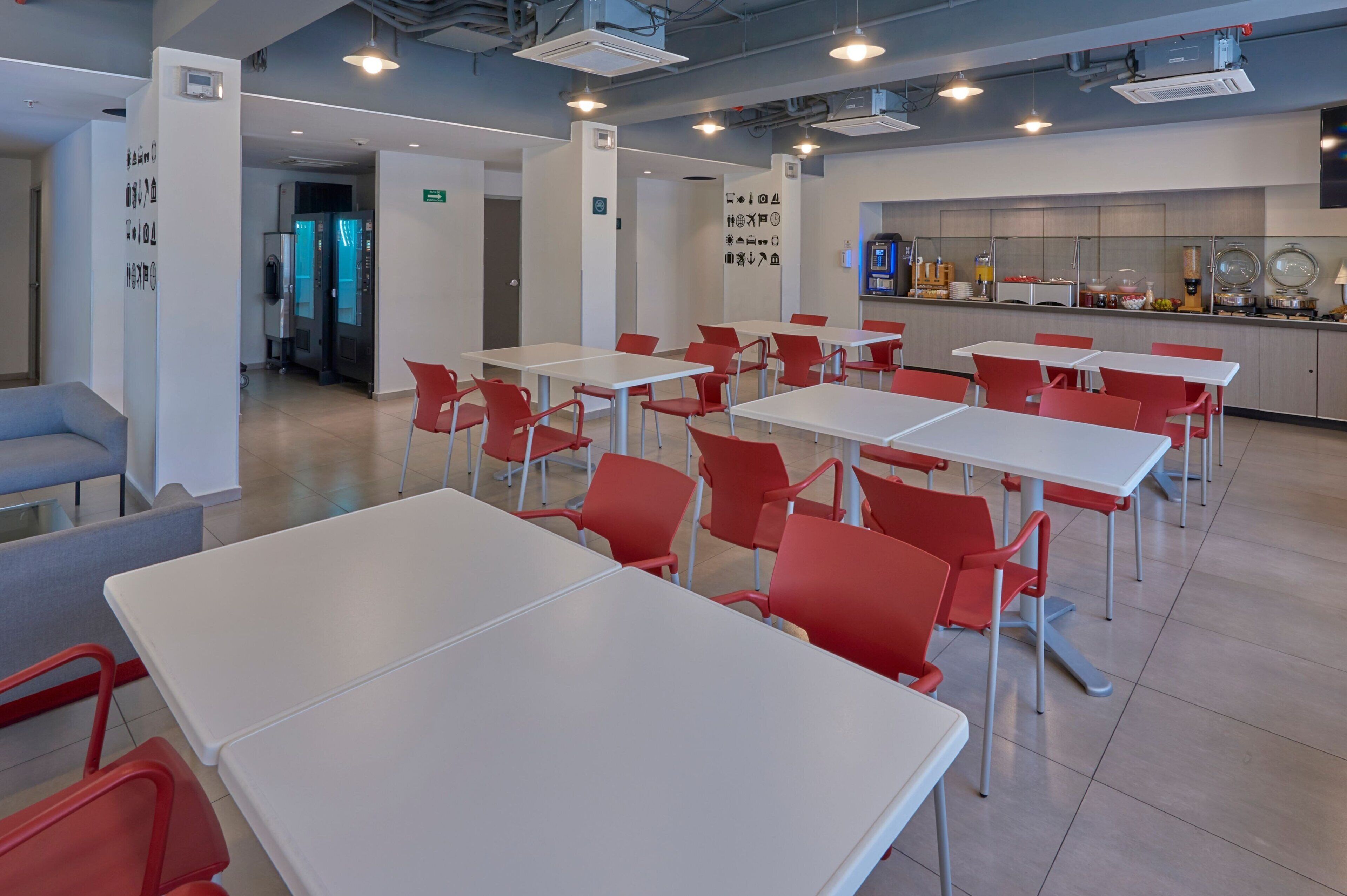 Un comedor moderno con mesas blancas y sillas rojas, una zona de cocina con diversos alimentos, y una puerta de salida visible.