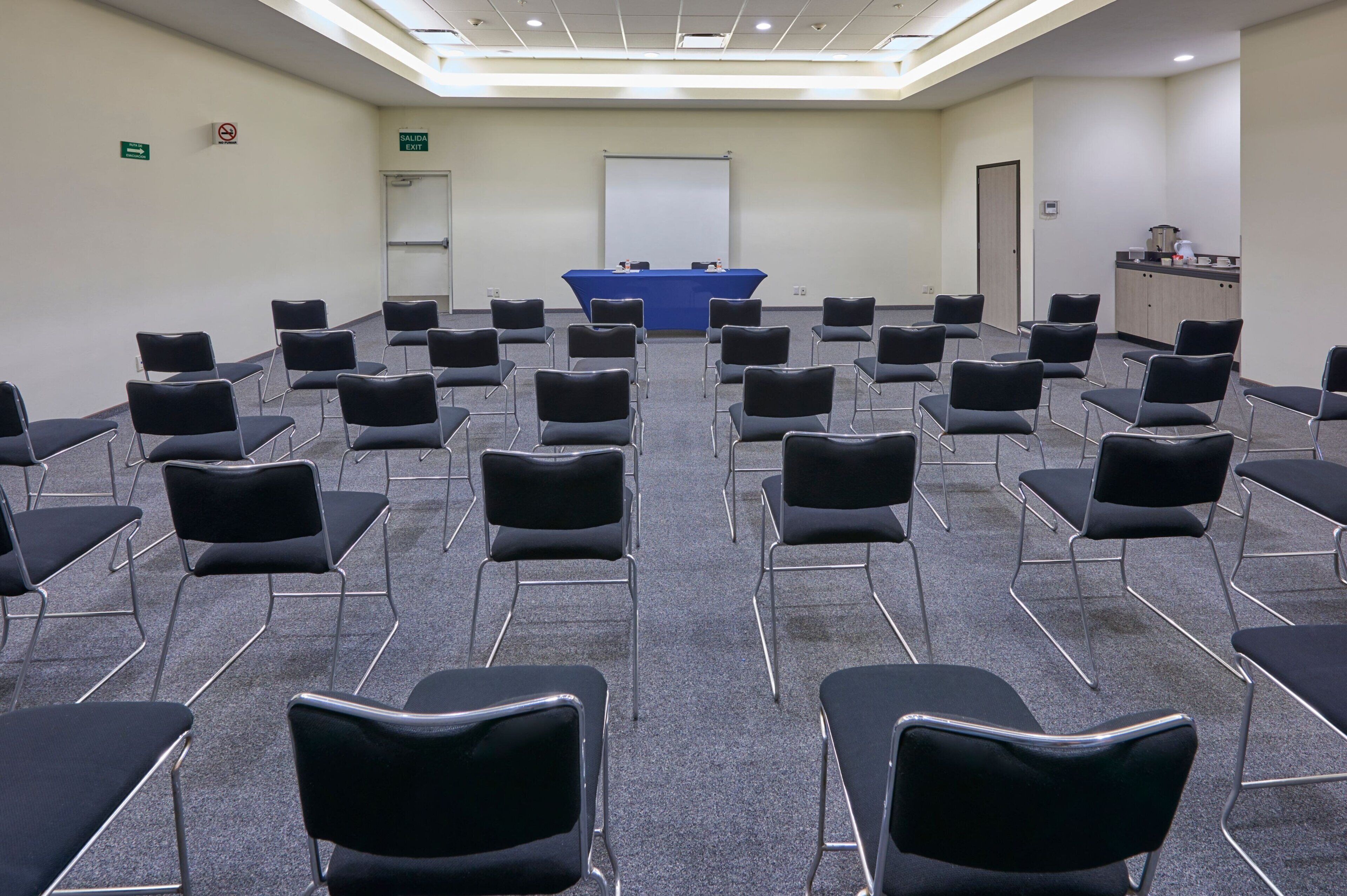 A conference room with rows of black chairs, a blue table with microphones, a whiteboard, and a kitchenette area.