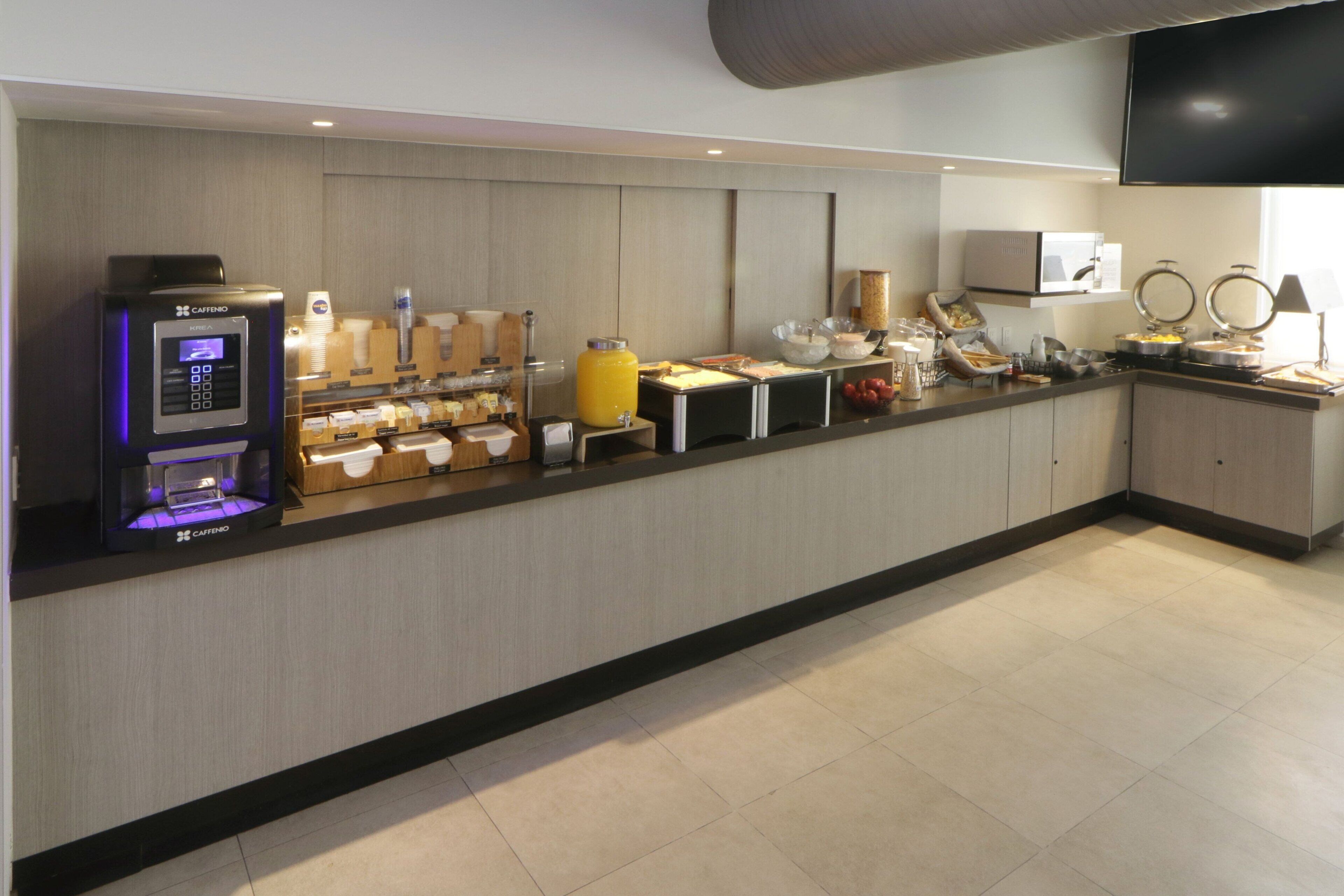 Une cuisine d’hôtel moderne dotée d’un espace de petit-déjeuner en libre-service, comprenant une machine à café, des fruits et divers produits alimentaires.