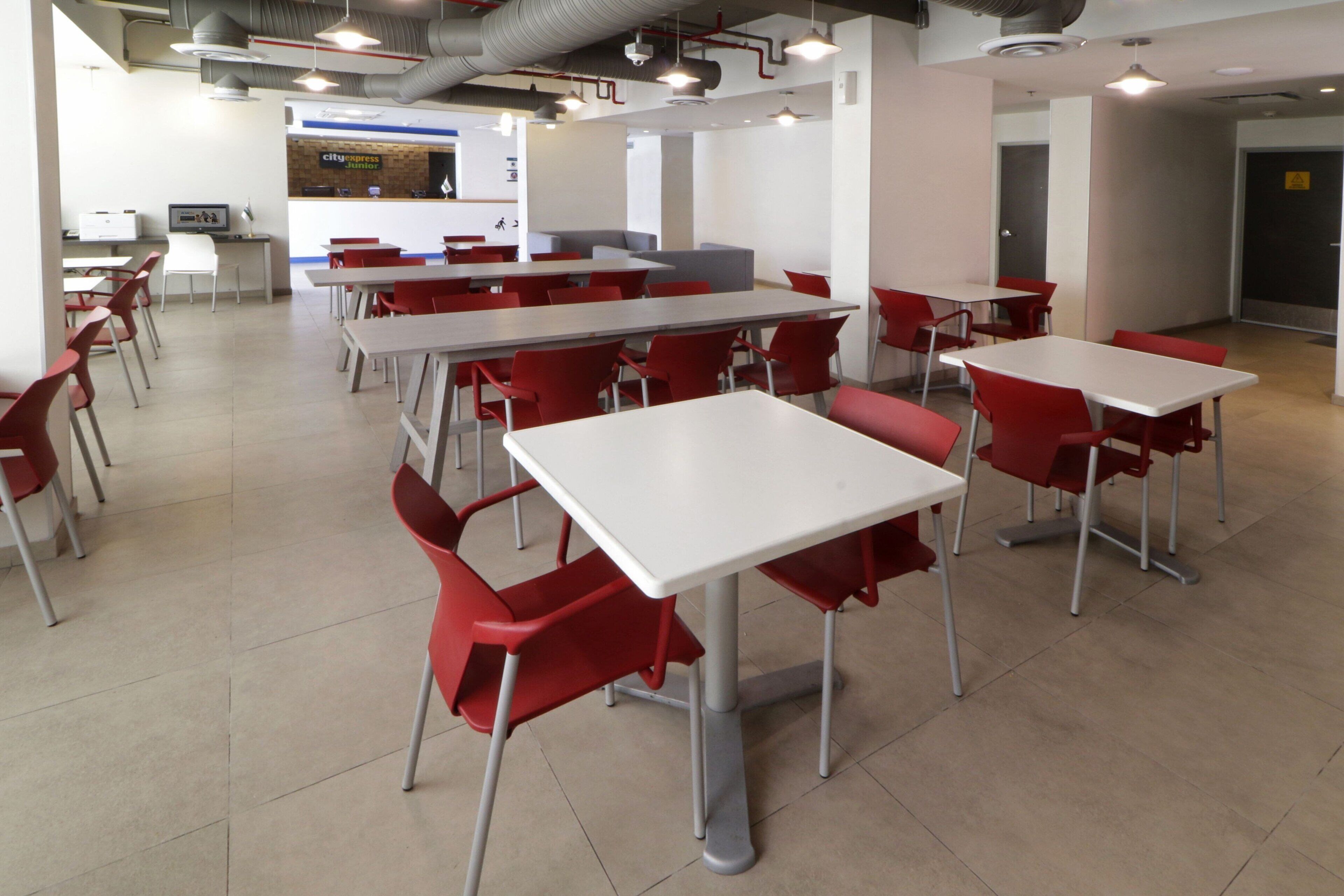 A spacious cafeteria with multiple tables and red chairs, a counter with a menu, and a seating area with a sofa.