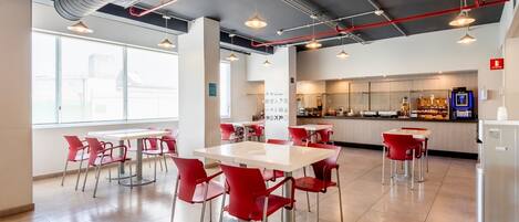 A modern cafeteria with white tables and red chairs, large windows, and a visible kitchen area.