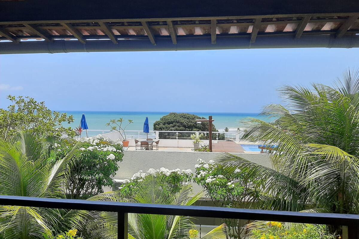 Vista da praia a partir de um espaço coberto, com guarda-sóis e assentos.