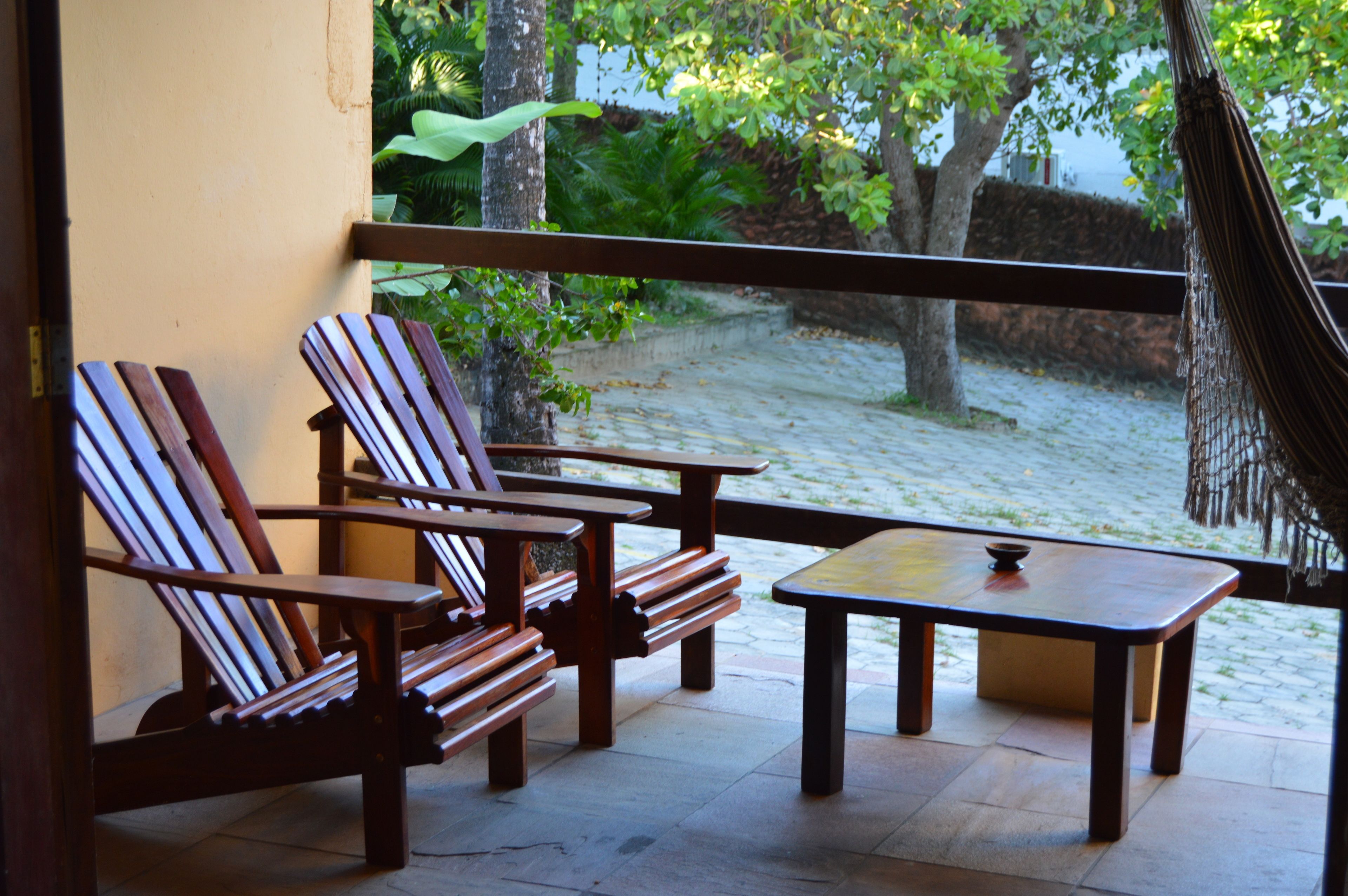 A balcony with wooden chairs and a table, overlooking a sandy area with trees and a hammock.