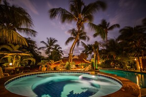 A swimming pool surrounded by palm trees and lounge chairs, with a thatched roof structure in the background.