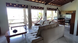 A spacious living area with a sofa, dining table, and chairs, a kitchen in the background, and large windows with a view of greenery. - Pousada Cavalo Marinho (Tibau do Sul)