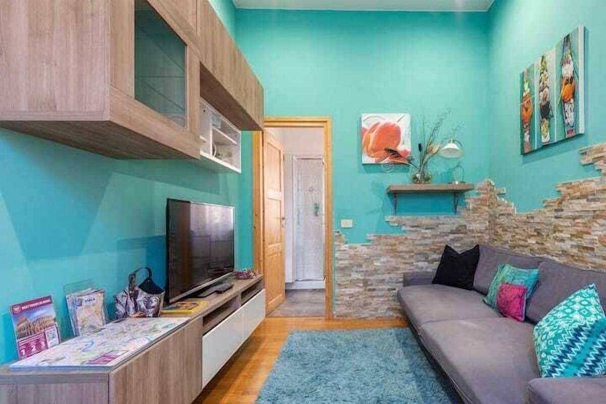 Ein Wohnzimmer mit türkisfarbenen Wänden, einem grauen Sofa, einem Fernseher und einer Wandgestaltung aus Stein.