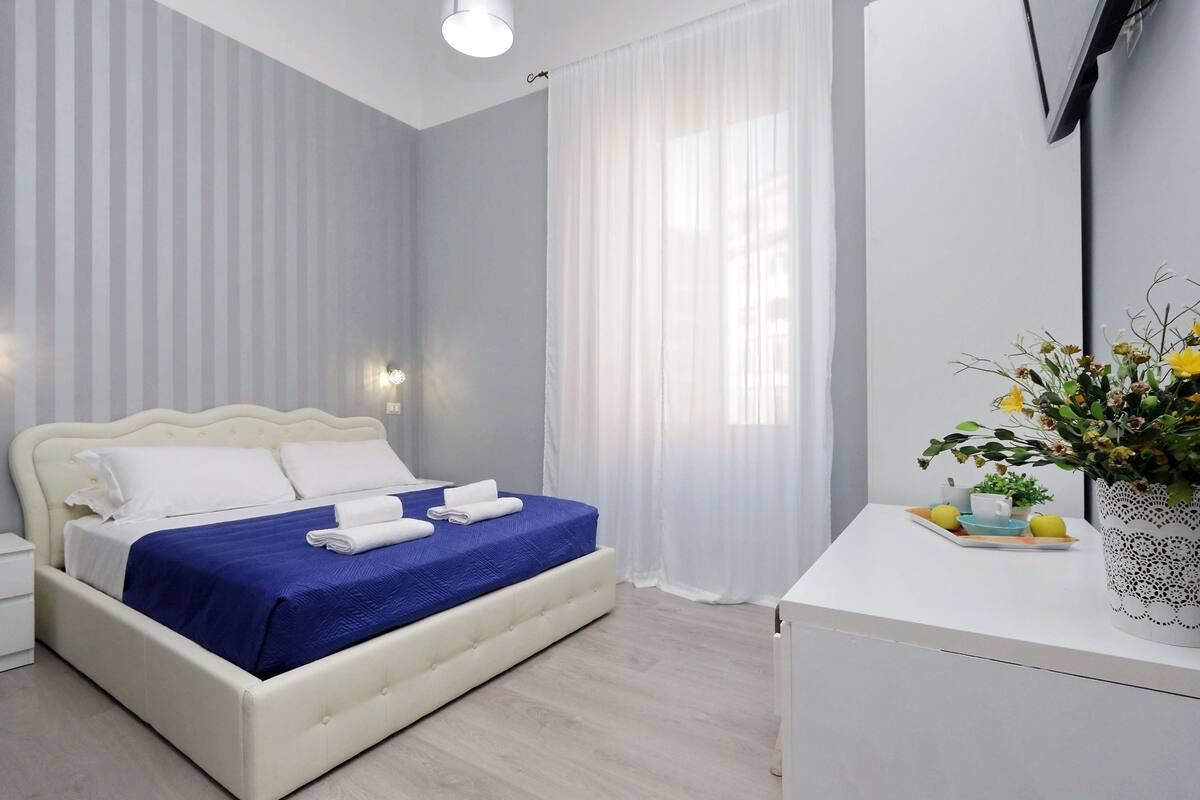 Ein ordentlich bezogenes Bett mit weißer und blauer Bettwäsche, ein Nachttisch mit Blumenvase und ein an der Wand befestigter Fernseher.