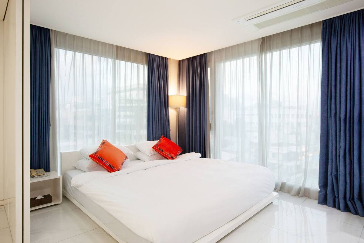 A modern hotel room with a large bed, a bedside table with a lamp, and a city view through sheer curtains.
