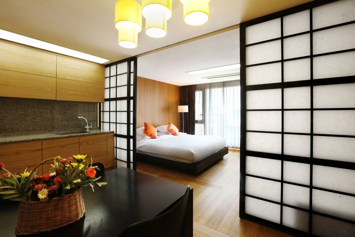 A modern hotel room with a wooden cabinet, a bed with orange pillows, a black-framed window, and a wall with a grid pattern.
