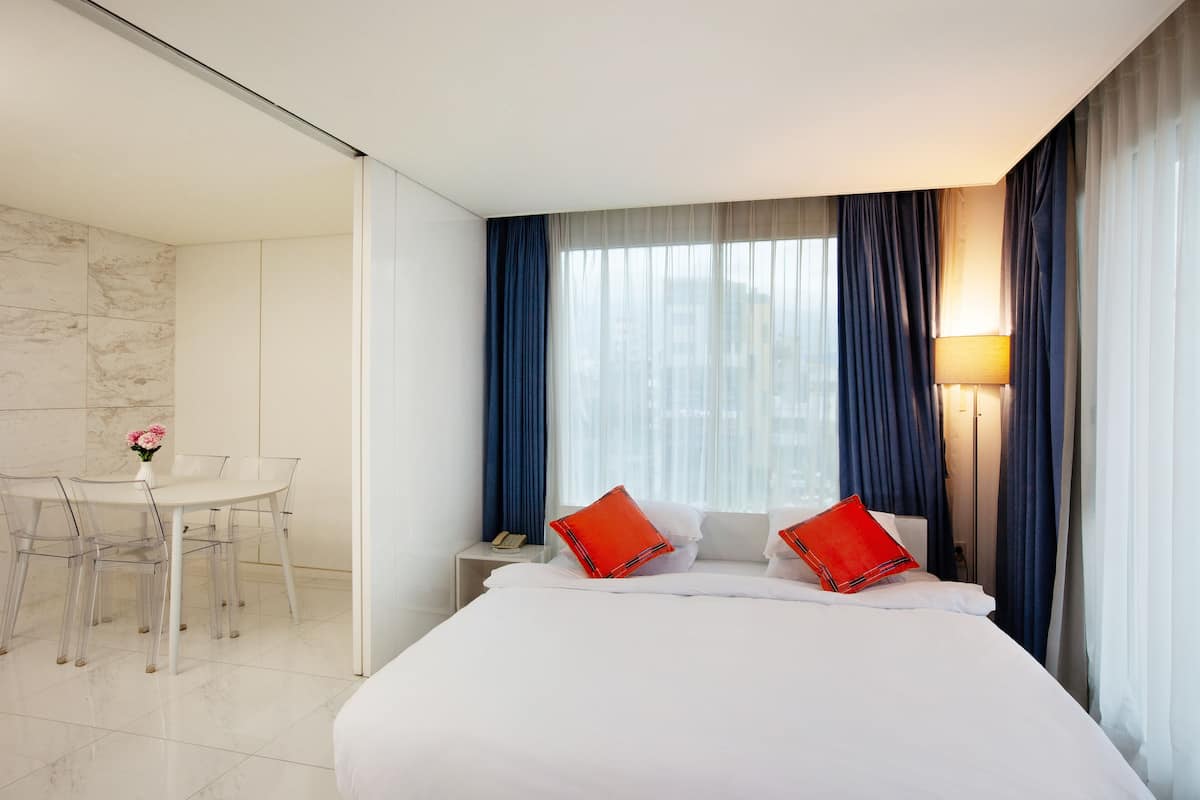 A hotel room with a bed, a bedside table with a lamp, a chair, and a small table with a vase of flowers.