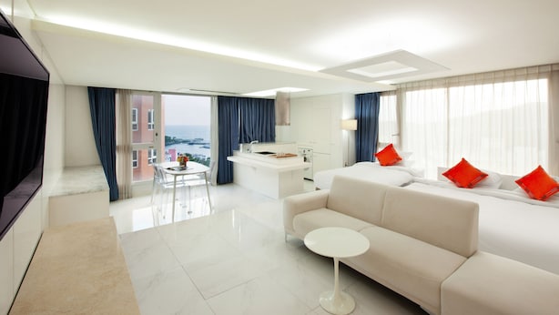 Une chambre d’hôtel moderne avec un canapé, une petite table ronde, un lit avec des oreillers rouges, un bureau avec une chaise et une vue sur la mer.