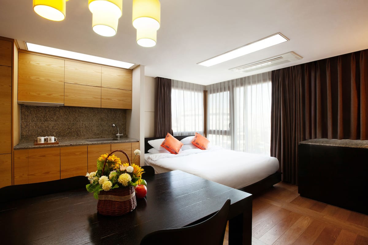 A modern hotel room with a wooden kitchenette, a large bed with orange pillows, a dining table with a basket of flowers, and a dark wooden floor.