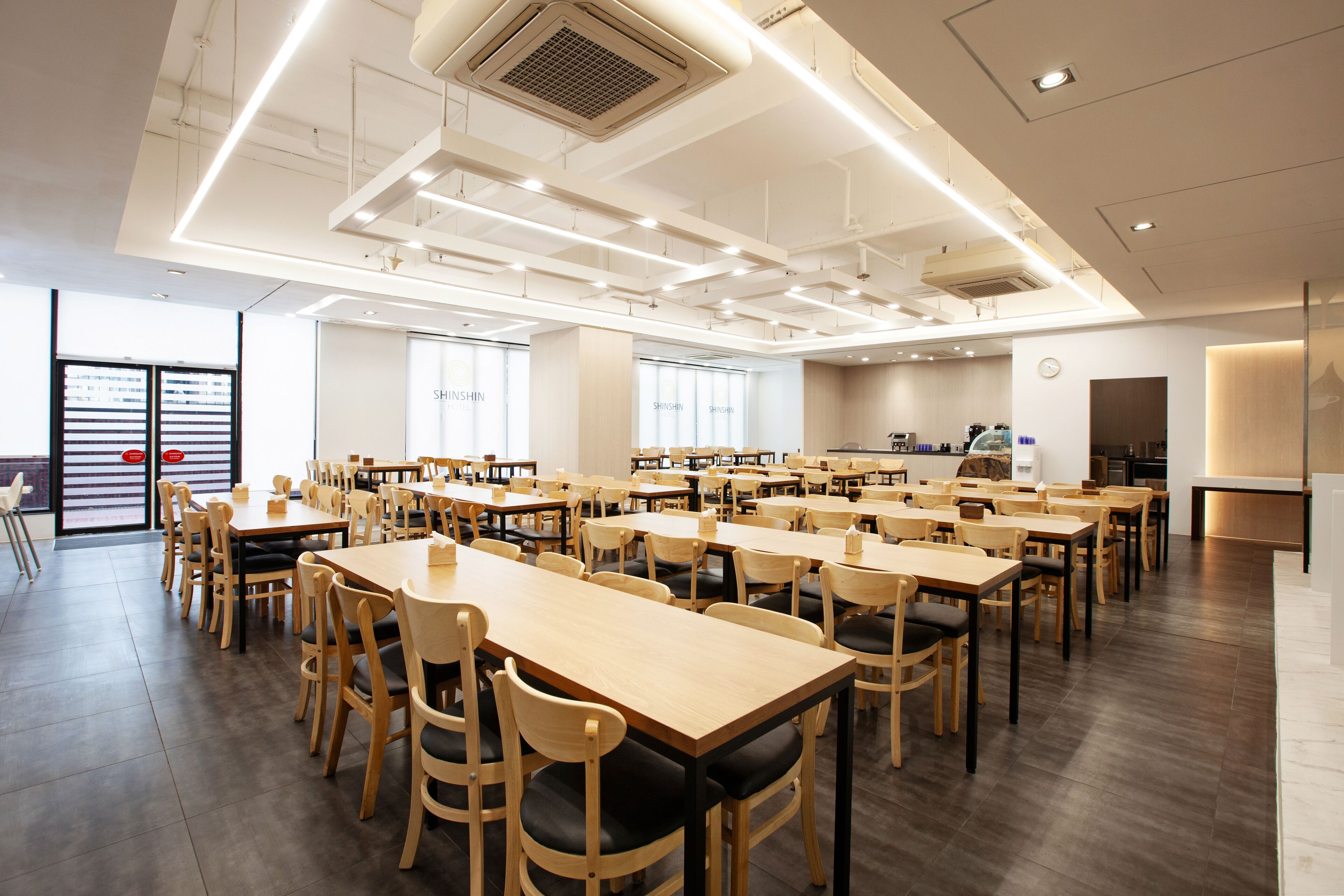 Un grand espace dédié à la salle à manger, avec plusieurs tables et chaises, une cuisine à l’arrière-plan et de grandes fenêtres qui laissent entrer la lumière naturelle.
