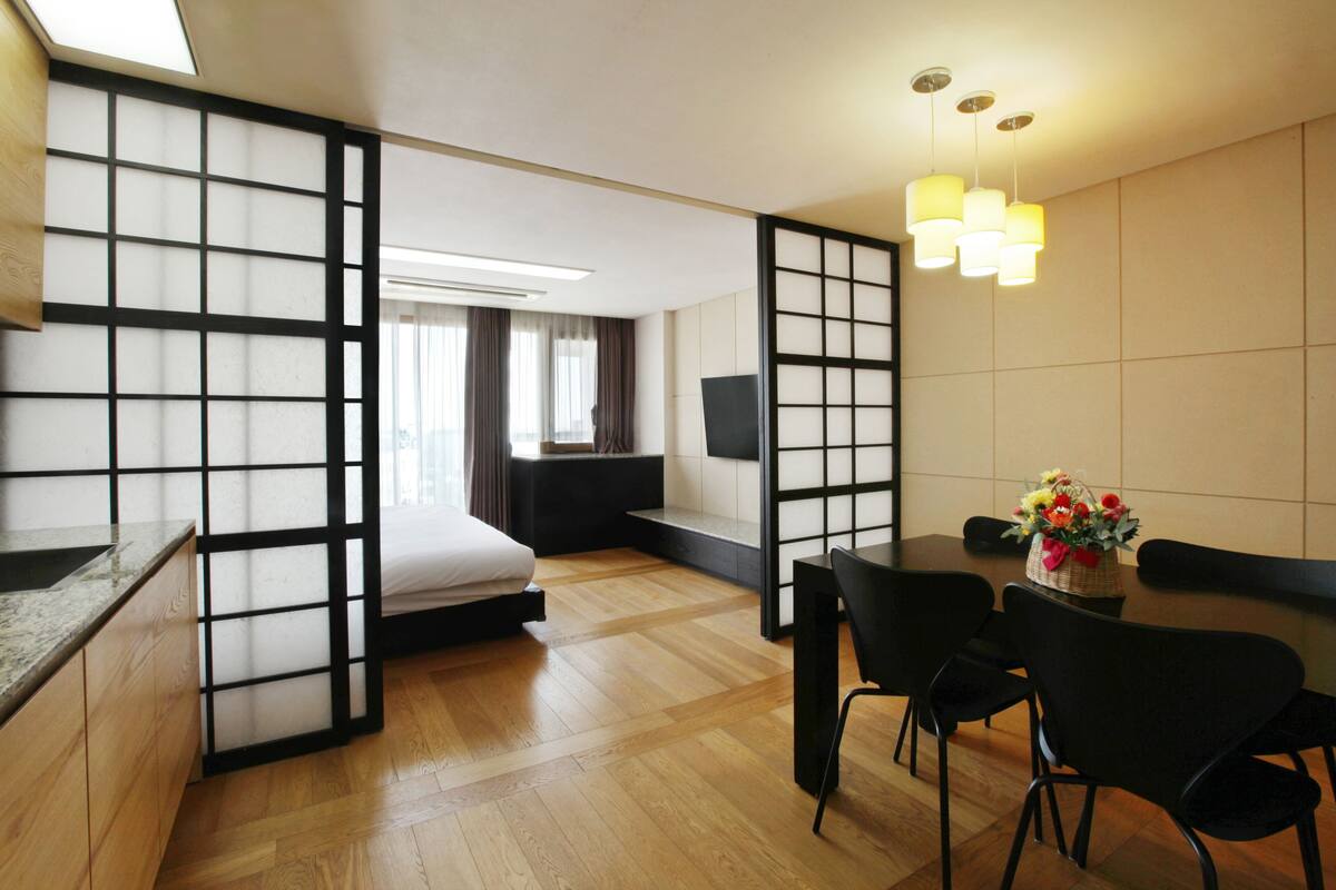 A modern hotel room with a wooden floor, a bed, a dining area with chairs, a table, and a decorative vase.