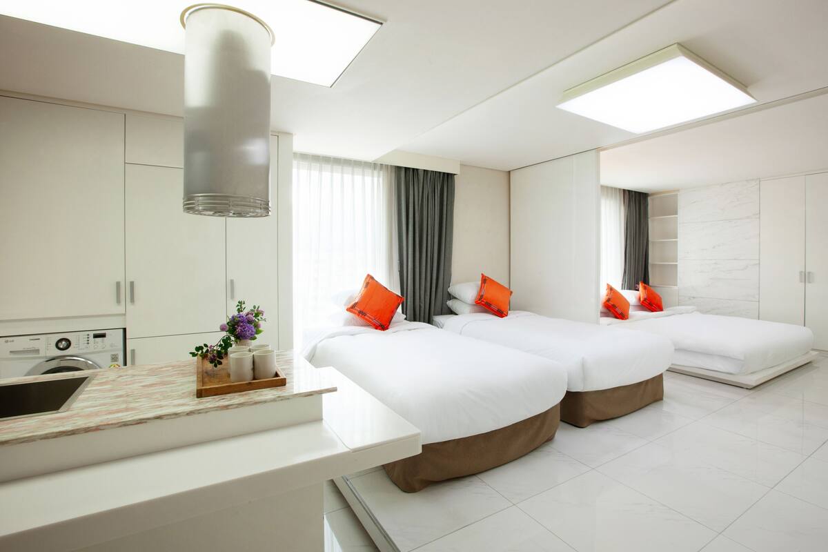 A modern hotel room with two beds, a kitchenette, and a bathroom.