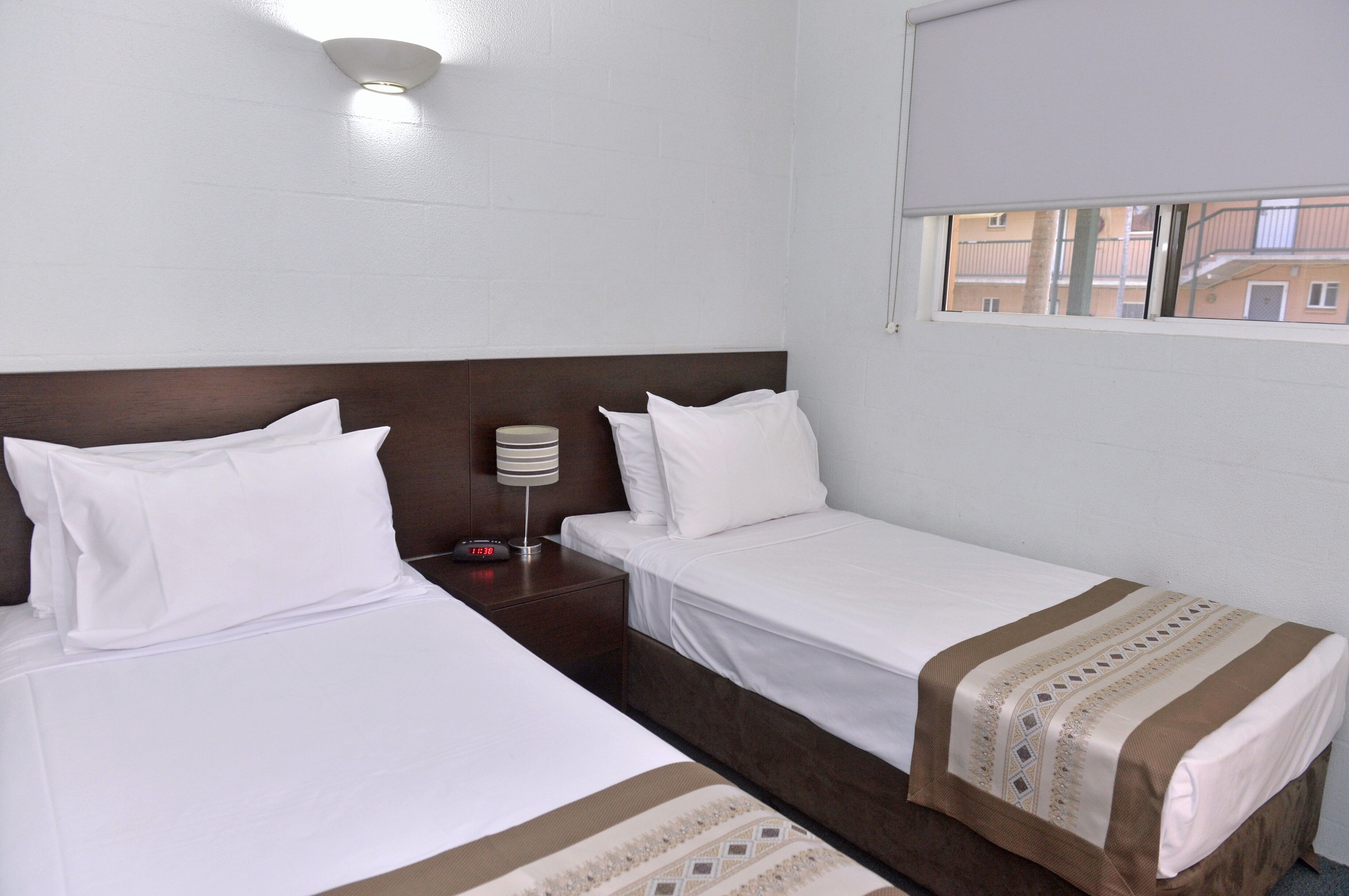 A hotel room with two single beds, a nightstand with a lamp, and a window with a view of buildings.
