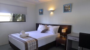 A neatly made bed with a white comforter and a brown headboard, a bedside table with a lamp, a desk with a chair, and a window with blinds. - Alatai Holiday Apartments (Darwin)
