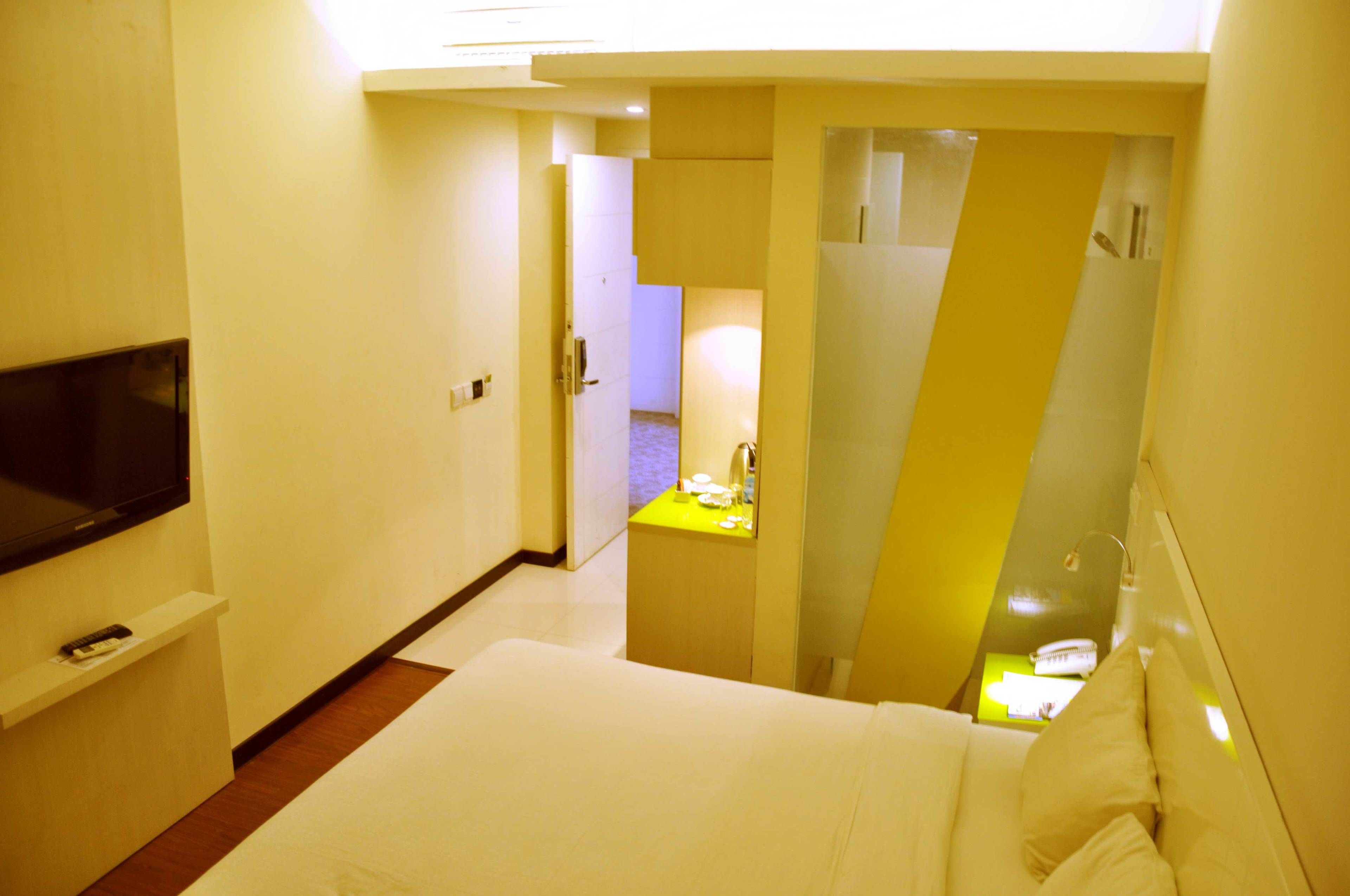 Habitación de hotel con cama, televisión montada en la pared, escritorio pequeño con lámpara y baño visible a través de una puerta.