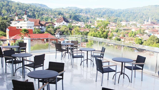 Une terrasse dotée de tables et de chaises rondes, offrant une vue sur une rivière et sur une ville avec ses bâtiments et sa verdure.