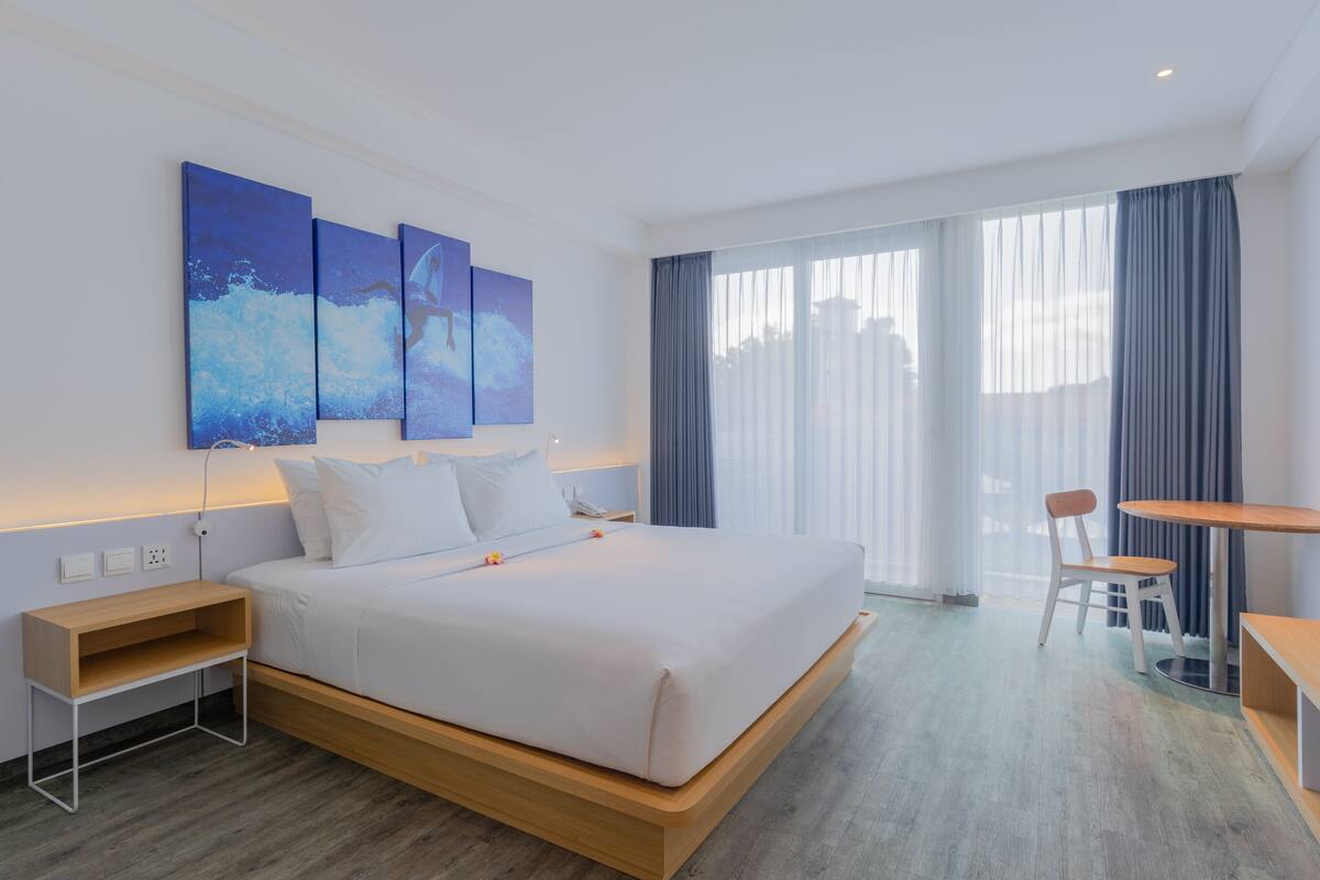 Une chambre d’hôtel moderne avec un grand lit, une table de chevet en bois, un bureau et une chaise. Il y a trois tableaux au mur et une fenêtre avec des rideaux.