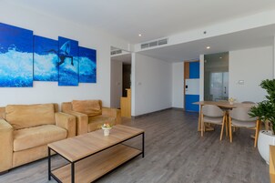 A modern living area with a sofa, coffee table, and a dining table with chairs. There is a large painting on the wall and a potted plant.