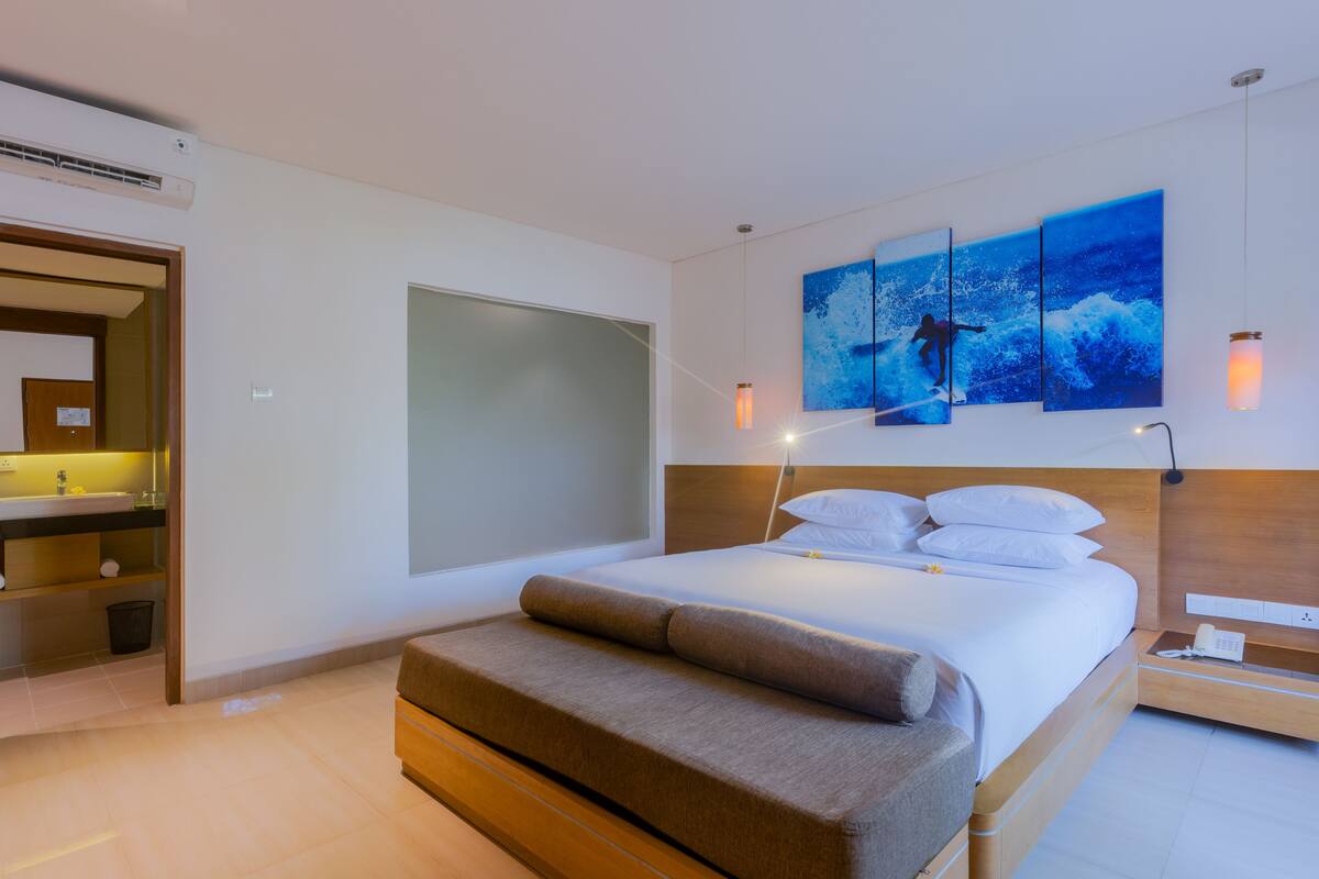 Une chambre d’hôtel moderne dotée d’un grand lit, d’une table de chevet, d’une commode, d’un climatiseur mural, d’une salle de bain avec un lavabo et un miroir, et d’une grande fenêtre.