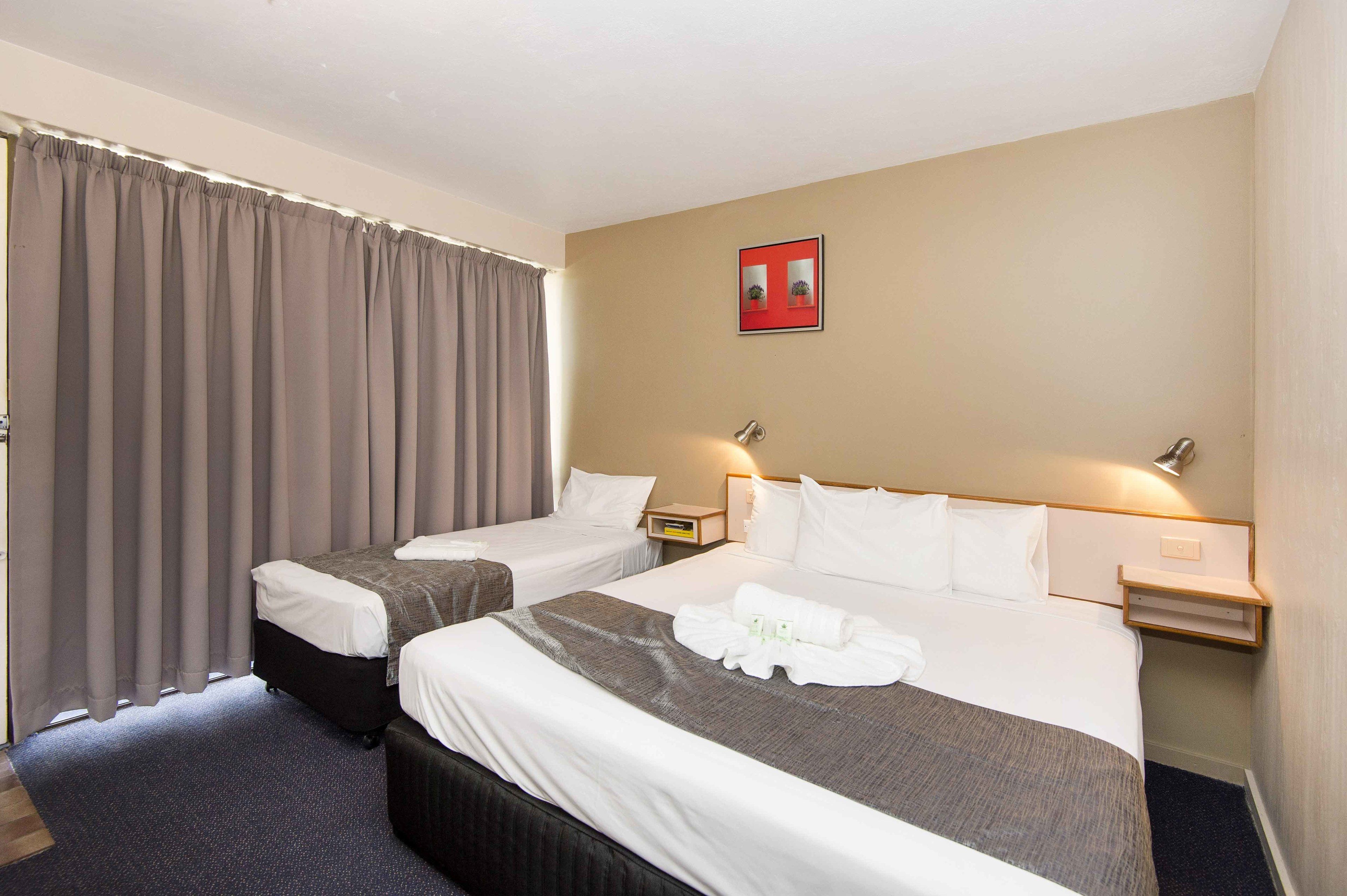 Ein Hotelzimmer mit zwei Betten, einem großen Fenster mit Vorhängen, einem Nachttisch mit Lampe und einem Wandbild.