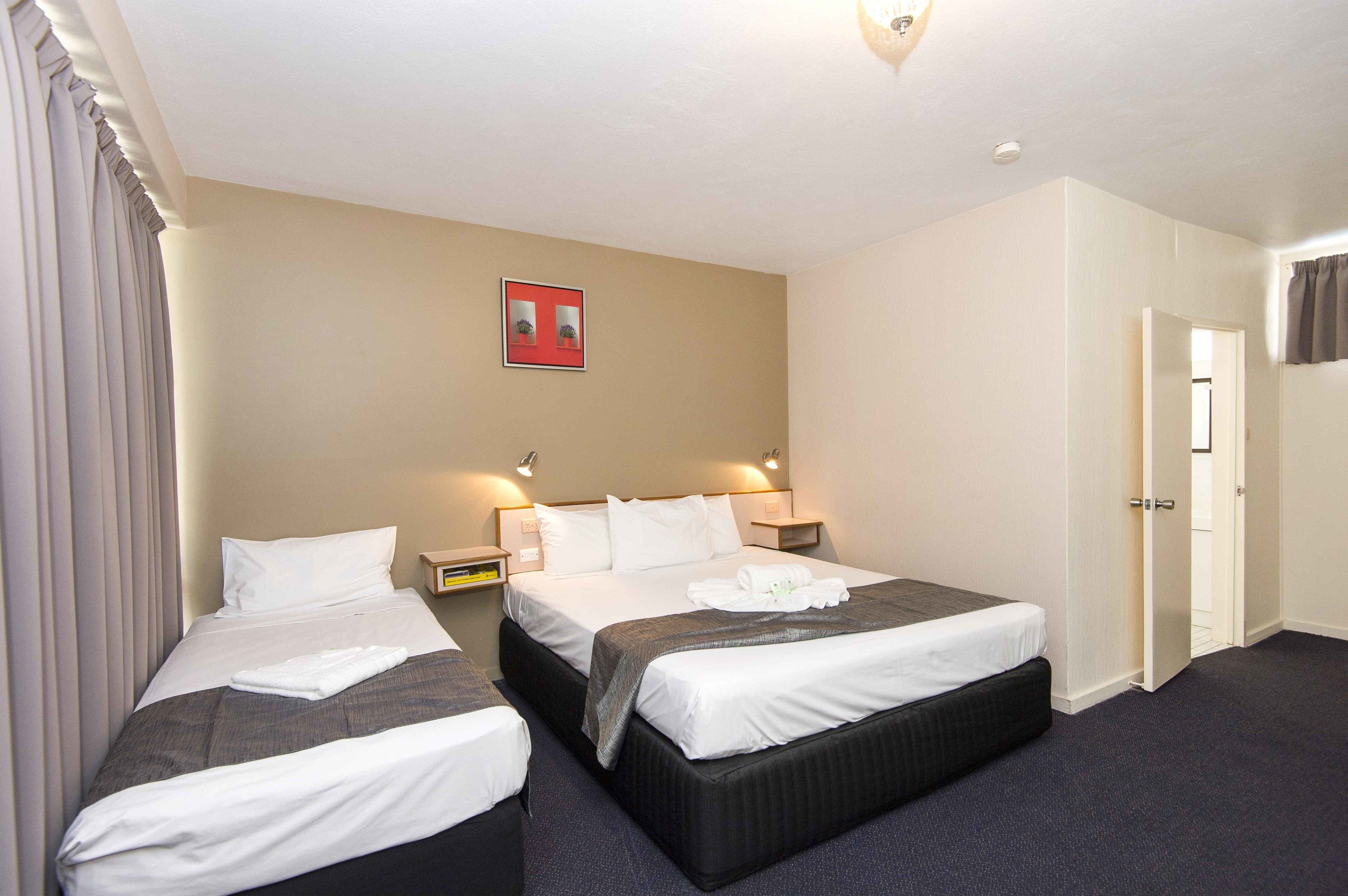A hotel room with two beds, a bedside table, a nightstand, a framed picture on the wall, and a door leading to another room.