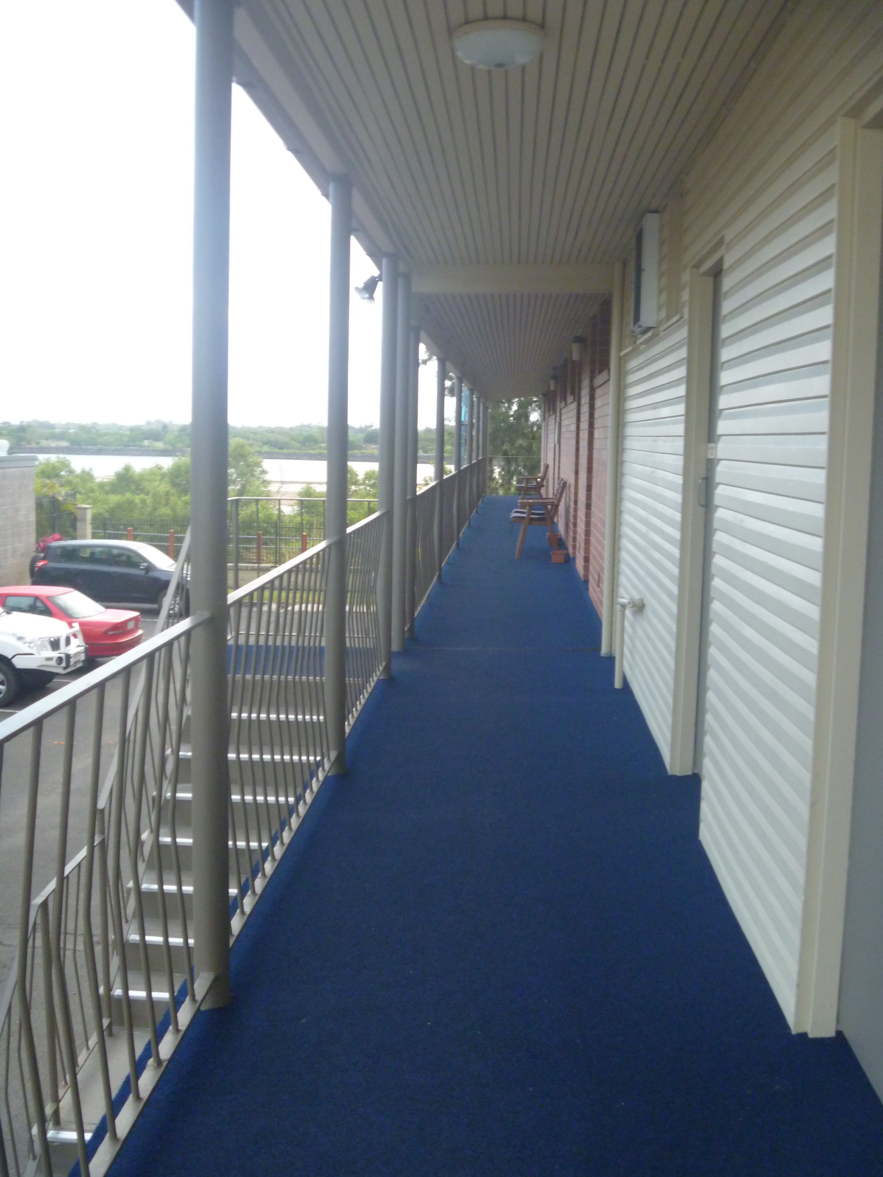 Un couloir couvert, doté d’une moquette bleue, d’une rambarde en métal et de volets fermés sur le côté droit.