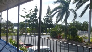 A balcony overlooking a parking area with a white car, palm trees, and a view of a body of water.
