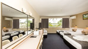 A hotel room with a large mirror, two beds, a desk with a computer, and a view of greenery outside.