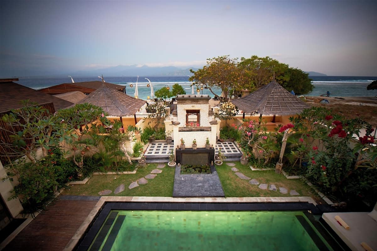 Zona de piscina con vista al mar, rodeada por un jardín con macetas y una pequeña construcción con techo de tejas.
