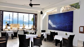 A restaurant with tables and chairs, a large painting on the wall, and a view of the sea. - Kokomo Resort Gili Trawangan (Gili Trawangan)